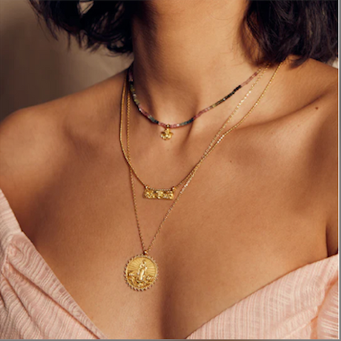 Close-up of a person wearing layered necklaces. The necklaces include a choker with colorful beads and a small pendant, a short chain with a gold bow pendant, and the Divine Mother Necklace featuring a round gold medallion depicting Our Lady of Guadalupe. The person is wearing a pink off-shoulder top.