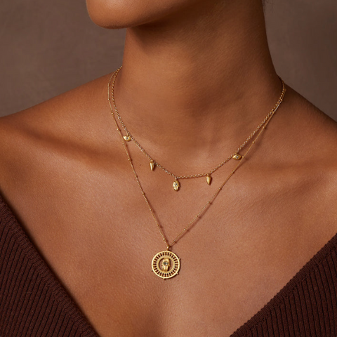 A person wearing two delicate gold necklaces. The shorter necklace features small, teardrop-shaped charms, while the longer one showcases the Palm of Protection Hamsa Coin Necklace with a round, ornate hamsa pendant. The individual is dressed in a deep brown, off-the-shoulder top.