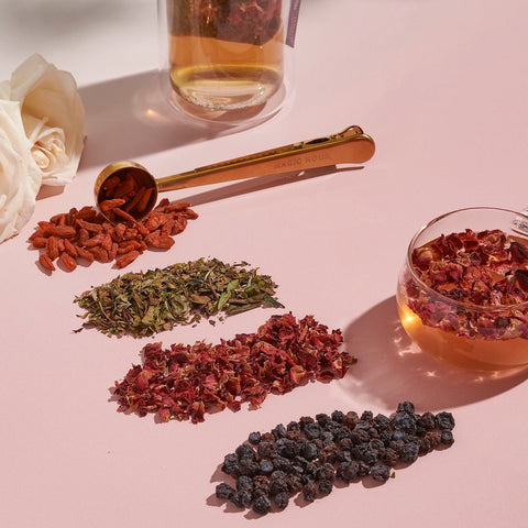 A pink surface displaying an assortment of dried herbs and berries in small piles, including goji berries, dried green leaves, rose petals, and dark berries. A gold measuring spoon and a clear cup with Passionfruit Green with Roses Iced Tea from Magic Hour are also present, along with a white rose in the background.