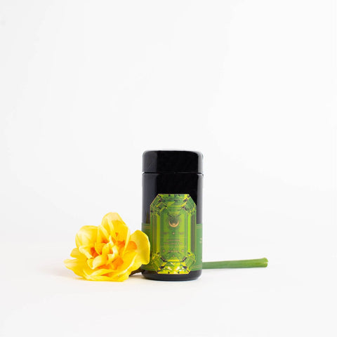 A refined black cylindrical container with a striking green label featuring the Magic Hour logo stands upright on a white background. To its left, a vibrant yellow flower with a green stem rests horizontally, infusing the minimalist scene with color and nature. The label offers a tantalizing hint of the refreshing aroma of organic mango within, announcing it as Peridot: Green Guayusa Oolong Mango-Peach Tea Case Packs.