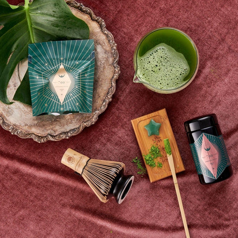 A flat lay image of a traditional matcha tea set, featuring the Chawan Matcha Bowl from Magic Hour. It includes a bamboo whisk, whisk holder, wooden tray with a star-shaped item, silver tray, matcha tea container, and green packaged box filled with loose leaf organic tea.