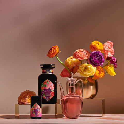 A vibrant still life composition features a golden vase with colorful flowers, a dark bottle and jar adorned with matching floral labels, and a glass decanter filled with pink liquid. The setup, which includes the immune-supporting *Pink Moment Pixie - Hibiscus Tangerine Dream Herbal Tea* by *Magic Hour*, is arranged on a beige background with golden platforms.