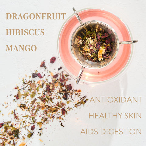 A clear teacup filled with Magic Hour's Pink Tourmaline: Organic Dragon Fruit Hibiscus White Tea sits on a white surface. Beside it, dried tea leaves are scattered. Text on the left reads "Dragonfruit, Hibiscus, Mango," and on the right, "Antioxidant, Healthy skin, Aids digestion." Enjoy immune support and promote heart & breast health with every sip.