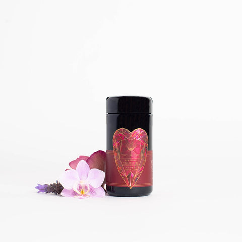 A black cylindrical container from the Magic Hour brand, adorned with a red label featuring a geometric heart design, stands beside three flowers: a white and pink orchid, a purple flower, and a red flower. The background is plain white. This Pink Tourmaline: Organic Dragon Fruit Hibiscus White Tea Case Pack promotes Heart & Breast Health while adding an elegant touch to your decor.