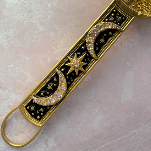 Here's a close-up of the 22-Carat Gold Plated Radiant Moon Gemstone Tea Strainer from Jaipur, India. This ornate, rectangular tea strainer features two crescent moons and a star adorned with pink cubic zirconia stones on a black background. Its gold-plated edge and detailed decorations create an elegant, celestial-themed design, all set against a light pink, textured background surface.