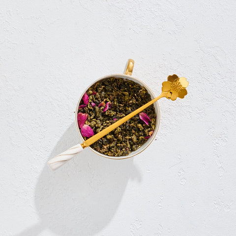 A cup filled with loose-leaf tea, adorned with pink flower petals, features the elegant Gold Floral Teaspoon resting across the top. The teaspoon, with its long gold stem and delicate white handle ending in a flower shape, is placed on a white textured surface.