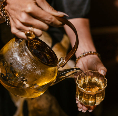 Close-up of a person pouring April 2024 Harvest - Grand Cru First Flush Organic "Spring Wonder" Samabeong Estate Darjeeling STGFOP1 from Magic Hour from a clear glass teapot into a small glass teacup. The teapot contains light-colored, organic tea from Samabeong Tea Garden, and steam can be seen rising. The person is wearing beaded bracelets and a ring on their finger.