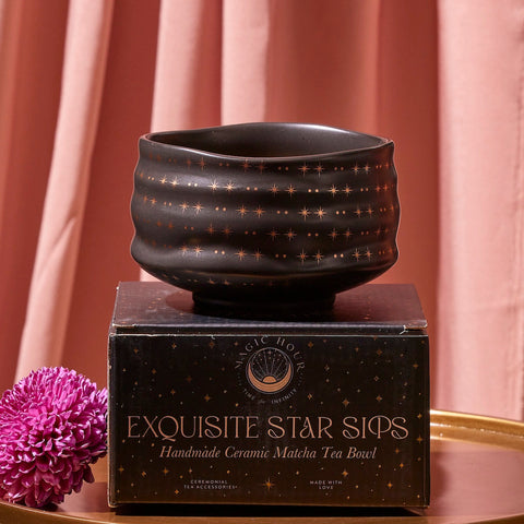 A dark, textured Matcha Bowl adorned with a star pattern rests on a black box labeled "Exquisite Star Sips: Handmade Ceramic Matcha Tea Bowl." Crafted using traditional methods and kiln firing, the background features soft pink drapes, and a purple chrysanthemum flower lies next to the box on a gold surface.