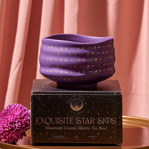 A handmade Matcha Bowl with a dark purple finish and gold star pattern, crafted by skilled artisans using traditional methods, sits atop a matching black and gold box labeled "Exquisite Star Sips." A pink flower and pink draped curtain serve as a backdrop on a golden surface.