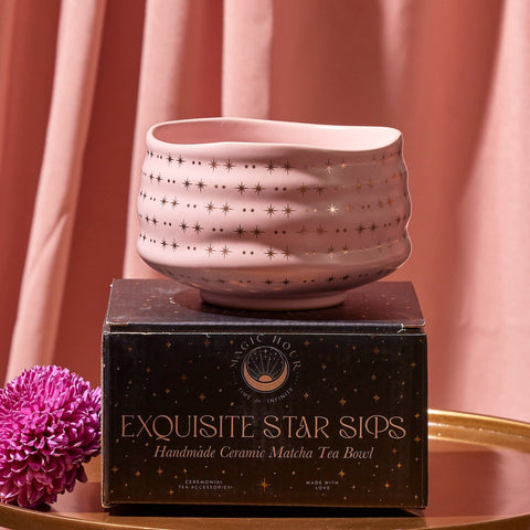 A pink Matcha Bowl with gold star patterns, crafted by skilled artisans using traditional methods, sits atop a black box labeled "Exquisite Star Sips: Handmade Ceramic Matcha Tea Bowl." The background is adorned with matching pink fabric, and a purple chrysanthemum flower is placed to the left of the box.