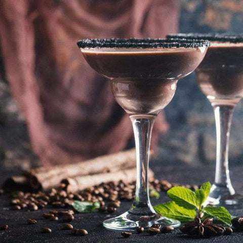 A close-up of two elegant cocktail glasses filled with a creamy chocolate beverage. The rims are coated with a dark substance, possibly sugar. Scattered coffee beans, cinnamon sticks, star anise, and mint leaves decorate the dark surface, evoking the enchanting aroma of Queen of the South: Delicious Cocoa Detox Tea by Magic Hour.