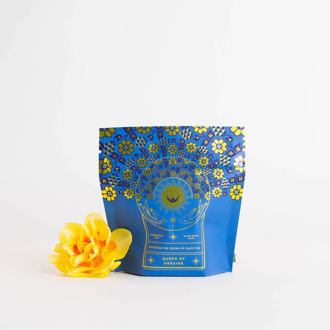 A blue tea packaging with gold floral patterns reads "Queen of Ukraine: Spring Blossom Tea, Magic Hour." The bag, adorned with a sun, moon, and stars design, sits beside a bright yellow flower on a plain white background. Experience the enchantment of Magic Hour's organic loose leaf blend.