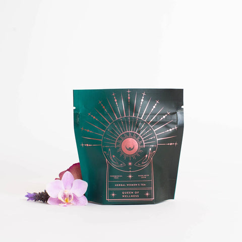 A teal and black package with a celestial design, labeled "Queen of Wellness: Women's Hormone Balancing Tea for PMS, Healthy Cycles & Menopause," offers an enchanting experience. Adorned with a golden sunburst and crescent moon, this Magic Hour tea is surrounded by an orchid bloom and lavender sprigs on a white background.