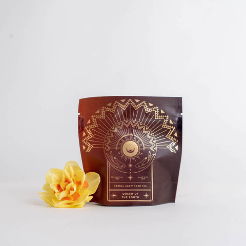 A sealed brown pouch labeled "Queen of the South: Delicious Cocoa Detox Tea - Magic Hour" with decorative gold designs, featuring a sun, crescent moon, and geometric patterns. Beside the Magic Hour pouch on a white surface lies a vibrant yellow flower.