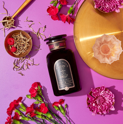 A black bottle labeled "Raspberry Earl White Tea" lies on a vibrant purple background, accompanied by a gold plate, scattered white tea leaves rich in antioxidants, red flowers, a small dish of raspberries, and a pink-lit candle in a rose-shaped holder.