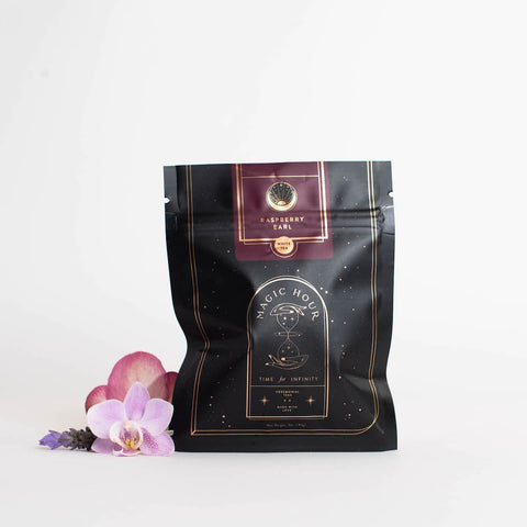 A black bag of Club Magic Hour Raspberry Earl White Tea with gold and red accents is displayed against a white background. The bag features elegant branding, including a logo and text. Bright purple and pink flowers are placed beside the bag for decoration, highlighting the loose leaf tea inside.