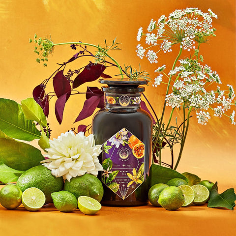 An elegant dark glass bottle labeled "Romance in Ravello" from Magic Hour is surrounded by fresh green limes, lime wedges, and assorted flowers against a vibrant yellow-orange background. This visually rich composition evokes the natural beauty and freshness of the Amalfi Coast, enhanced by hints of chocolate, passion fruit, and rose cream.