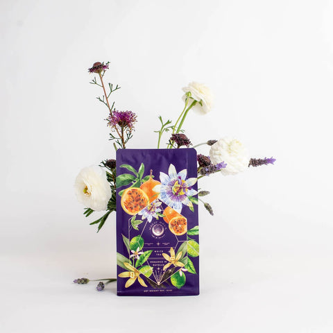 A beautifully designed coffee bag in a striking purple hue, adorned with vibrant illustrations of fruits and flowers, including a blend of passion fruit, sits gracefully in front of an exquisite floral arrangement featuring white roses, lavender, and other blossoms against a plain white background. This stunning display evokes the charm of mornings on the Amalfi Coast and perfectly showcases the Romance in Ravello - Chocolate-Passionfruit-Rose-Cream White Tea by Magic Hour.