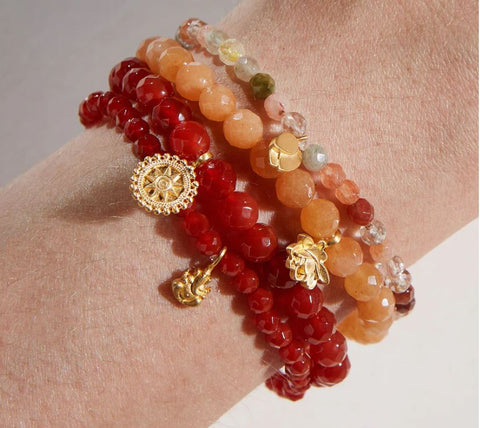 Wrist adorned with the **Red Carnelian Mandala Bracelet**, featuring multiple beaded strands in vibrant shades of red, orange, and translucent white. This healing jewelry piece also boasts gold decorative charms, including a compass, a horn, and a leaf. The beads come in various sizes, creating a colorful and layered look.
