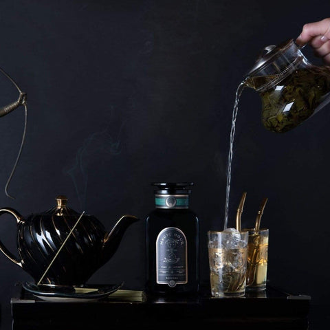 A hand pours **Magic Hour Renewal: Peach-Goji-Rose Oolong Tea** from a glass kettle into a glass filled with ice beside a black teapot. Behind the glass, an ornate bottle is displayed against a dark background. Two more glasses with golden straws rest nearby, creating an elegant drink setup.