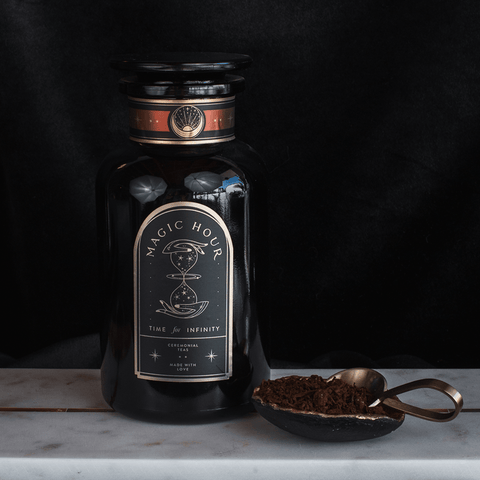 A dark glass jar labeled "Club Magic Hour" rests on a marble surface against a black background. Beside the jar, a brass spoon lies on a small dish filled with loose tea leaves, including Rooibos. The label showcases an hourglass along with the words "Time for Infinity" and "Hand-Crafted Tea.