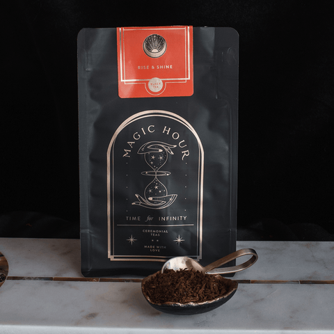 A package of "Rise & Shine Herbal Tea Case Packs" from Club Magic Hour sits on a marble surface. The black bag, adorned with gold and red detailing, is accompanied by a spoonful of rooibos tea leaves in front. The design features a celestial theme and reads "Time for Infinity.