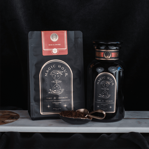 A photograph of two Club Magic Hour tea products on a dark background. To the left is a black and red "Rise & Shine Herbal Tea Case Packs" pouch with a celestial design. To the right, there's a matching black tea jar. A golden spoon with chaga mushroom-infused tea leaves is placed in front of the jar.