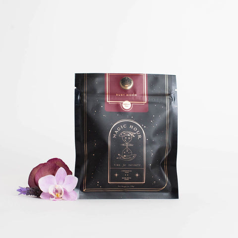 A black and gold pouch labeled "Ruby Moon™ : Hibiscus Elderberry Tea" rests against a white background. The pouch, containing organic loose leaf tea by Club Magic Hour, features galaxy-themed designs. Beside it lies a small arrangement of purple and white orchids.