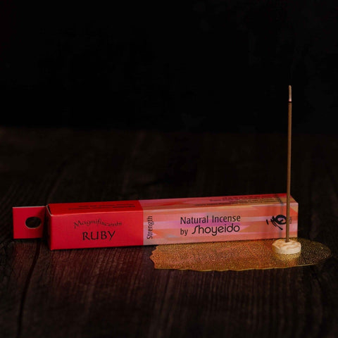 A stick of burning sandalwood incense stands upright in a small holder on a dark wooden surface. Positioned next to it is a red and pink box labeled "Natural Incense : Ruby" by Shoyeido. A decorative golden mesh lies beneath the holder.