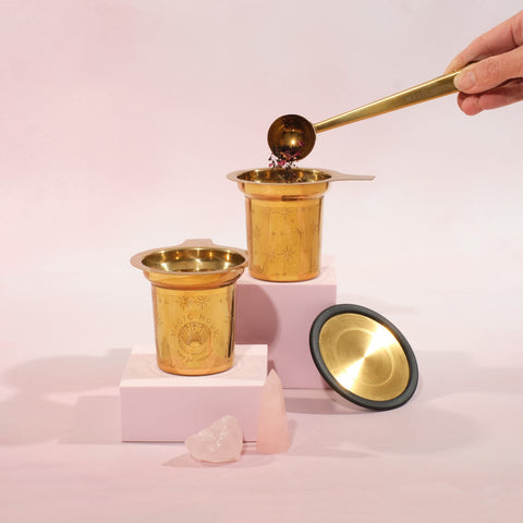 A decorative gold cup holds organic loose leaf tea, with a hand using a gold scoop to add more. Another identical gold cup and a lid sit nearby on two white platforms. Two small pink crystals are placed in the foreground against a soft pink background, creating the perfect Magic Hour Steep The Stars - Artisan Tea Strainer setting.