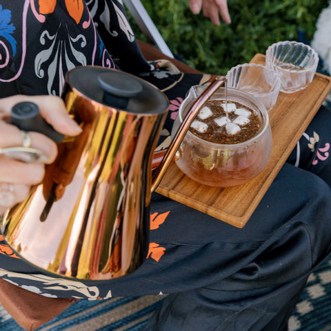 A copper tea kettle pouring hot water into a glass teapot filled with S'mores Herbal Dessert Loose Leaf Wellness Tea on a Wooden tea tray.