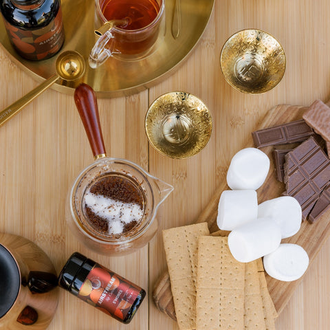 A top-down view of a wooden table set for s'mores preparation. Ingredients include marshmallows, graham crackers, and chocolate bars on a wooden board. A cup of S'mores Tea by Magic Hour, tea-making utensils with loose leaf tea, a jar of spice, and two ornate golden cups are also present.