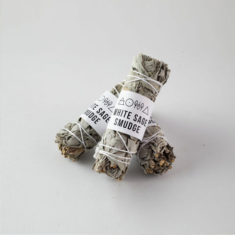 Three bundles of the "White Sage Smudge" product, tied with white string, rest on a white surface. Each sustainably sourced bundle features a label reading "White Sage Smudge," adorned with various small symbols.