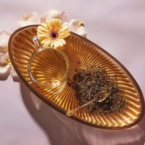 A glass teacup filled with Magic Hour's April 2024 Harvest - Grand Cru First Flush Organic "Spring Wonder" Samabeong Estate Darjeeling STGFOP1 on a golden oval tray, next to a small heap of loose organic tea leaves and a golden spoon. The tray is adorned with a single yellow gerbera daisy and white rose petals, all set against a soft pink background.