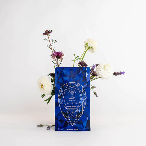 A blue geometric-patterned tea bag labeled "Sapphire - Apple-Plum-Spice White Tea Case Packs" from Magic Hour is centered against a white background. The tea bag is surrounded by an arrangement of flowers, including white roses and purple blooms, emerging from behind it.