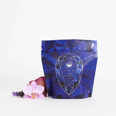 A blue, geometrically designed pouch labeled "Sapphire - Apple-Plum-Spice White Tea Case Packs" from Magic Hour is displayed against a white background. Beside the pouch, there is a light pink orchid and a small purple flower, adding a touch of natural elegance to the image of this calming grounding tea.