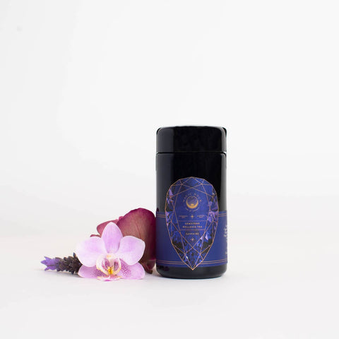 The "Sapphire - Apple-Plum-Spice White Tea Case Packs" by Magic Hour, housed in a black cylindrical container with a purple geometric label featuring gold details and text, stands next to a pink orchid flower and a red rose petal, all placed on a white surface with a white background.