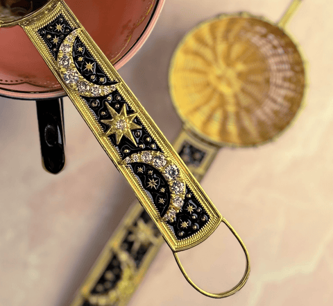 Close-up of intricately designed gold and black coffee pot handles adorned with crescent moons, stars, and Cubic Zirconia stones. One 22-Carat Gold Plated handle is attached to a copper pot, while the Radiant Moon Gemstone Tea Strainer from Jaipur, India lies next to a brass tea basket on a light-colored surface.