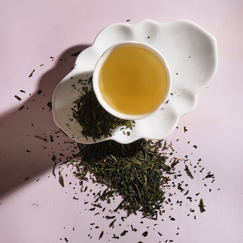 A white ceramic teacup filled with light Sencha Kyoto Green Tea rests on a uniquely shaped white saucer. Loose green tea leaves, rich in antioxidants and known to boost metabolism, are scattered around the saucer and teacup on a soft pink background. The image is well-lit, highlighting the texture of the leaves and the tea's color.