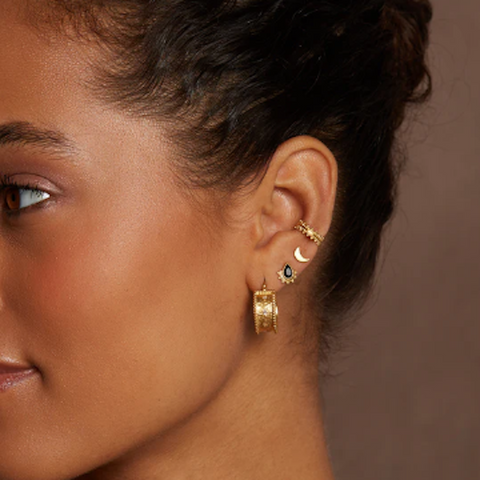 A close-up of a personalized ear stack adorned with multiple gold earrings and the Magic Hour Shine On Beaded Ear Cuff, including a small hoop with a sun charm, a crescent moon stud, a small bi-color stud, and a larger textured hoop earring at the lobe. The background is plain brown.