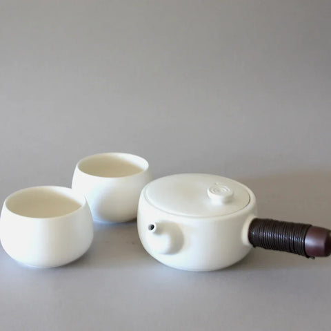 A minimalist Side Handle Tea Set by Eastern Elm, featuring a white ceramic teapot with a cylindrical brown handle and two round tea cups placed beside it on a plain, light grey background. The set combines traditional craftsmanship with modern, sleek design.