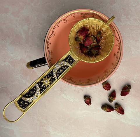 A 22-Carat Gold Plated Radiant Moon Gemstone Tea Strainer from Jaipur, India, adorned with intricate celestial designs and cubic zirconia stones, is placed over an empty pink teacup with a matching saucer. The strainer contains dried rosebuds, while more rosebuds are scattered around the teacup on a light pink surface.