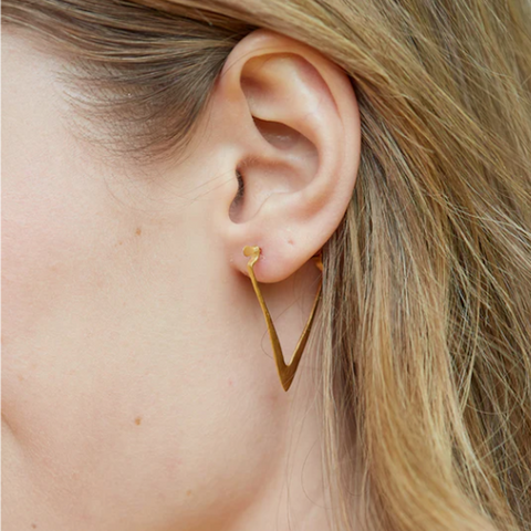 Close-up of a woman's ear wearing Magic Hour's Simple Strength Earrings, a delicate, 18k gold V-shaped earring. The modern silhouette dangles slightly below the ear lobe, while her light brown hair partially covers the ear. The background is blurred, keeping the focus on the ear and earring.