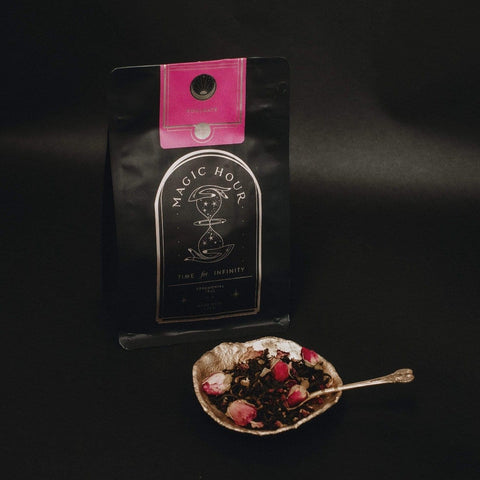 A package of Magic Hour Tea "Soulmate: Chocolate-Raspberry-Rose Black Tea for Finding & Celebrating Love" with a pink label sits against a dark background. In front, a decorative bowl brims with organic tea leaves and dried rosebuds, while a small spoon rests on the bowl.