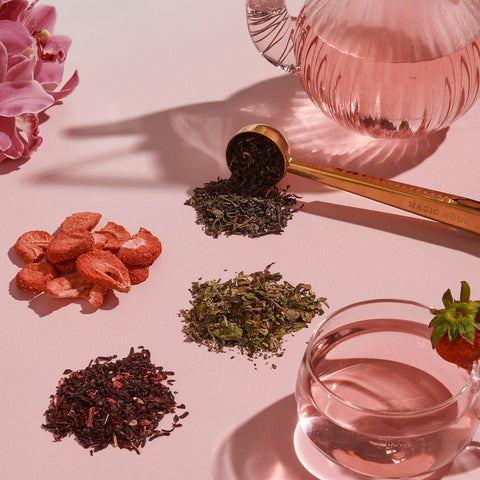 A pink tea set up with loose Strawberry Shortcake Iced Tea with Hibiscus leaves from Magic Hour and dried strawberries on display. A glass teapot and teacup filled with pink liquid are complemented by a gold tea scoop. Pink flowers decorate the background, adding an elegant touch to the arrangement.