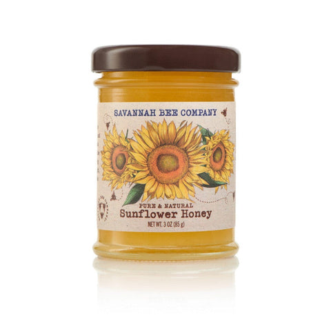 A jar of Savannah Bee Co. Honey - Sunflower, pure and natural, features a label adorned with images of three sunflowers and bees, evoking the beauty of sunflower fields in Ukraine and Romania. The jar comes with a dark brown lid and holds 3 ounces (85 grams) of delicious honey.