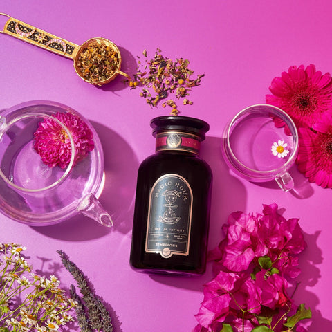 A beautiful display featuring a jar of Symbeeosis: Beautifying Immunitea for the Queen Bee on a vibrant pink background. Surrounding the jar are a transparent teapot, two transparent cups, a spoon with loose leaf tea, and assorted colorful flowers. This serene setup invites you to start your wellness journey with health-boosting ingredients.