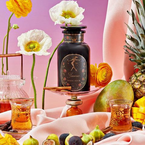 A decorative bottle labeled "Taurus: Tea of Venusian Garden Delights" from Magic Hour is surrounded by vibrant yellow and white flowers, fresh fruit including pineapple, mango, and figs, with a pink and purple gradient background. Two ornate glasses with amber liquid from the Magic Hour collection are placed in the foreground.