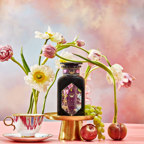 A bottle of "Breakfast in Paris-Oolong Black Tea Scented with Fruits & Flowers of Secret Parisian Gardens" is elegantly showcased on a gold stand, accompanied by flowers, fruits, and a teacup with a saucer. The background captures the essence of secret Parisian gardens, featuring a pastel gradient of soft pink, blue, and yellow tones.