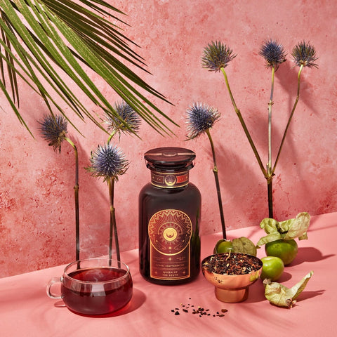 A dark bottle with a sun emblem stands on a pink surface surrounded by exotic flowers, green fruits, a teacup filled with a dark red liquid, and a bowl of organic loose leaf tea. A large green leaf dangles from above, set against a textured pink background. Magic Hour Queen of the South: Delicious Cocoa Detox Tea awaits your senses.